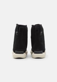 Botas de tobillo de ante negro con un forro suave, que presentan un logo en el tacón y una suela de goma blanca en contraste. Diseño sencillo y moderno.