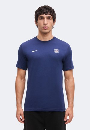 Uomo in piedi rivolto in avanti, indossa una maglietta Nike blu con il logo del Paris Saint-Germain sul petto e pantaloni neri.