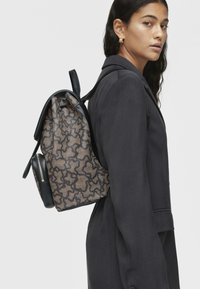 Zaino con dettagli in pelle nera, caratterizzato da un motivo beige texturizzato. Ha una parte superiore arrotondata e spallacci sottili, indossato su una spalla.