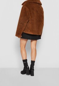 Bruine jas van nep bont met een brede kraag, gecombineerd met een zwarte minirok en stevige zwarte laarzen met hak, waarbij geribbelde sokdetails zichtbaar zijn.