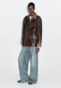 Veste en cuir marron avec un col couleur beige, poches avant et coutures contrastées, associée à un jean large bleu clair et des chaussures foncées.