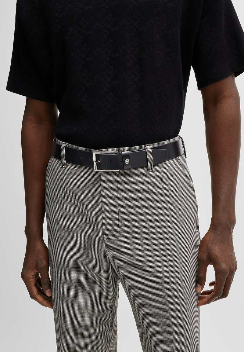 Ceinture en cuir noir avec une boucle en argent, portée avec un pantalon gris à carreaux et un t-shirt noir texturé. Accent sur le contraste des matériaux et des couleurs.