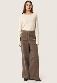 Young woman standing, wearing a cream long-sleeve top, high-waisted wide-leg brown corduroy pants, and black shoes, on a plain light background.
