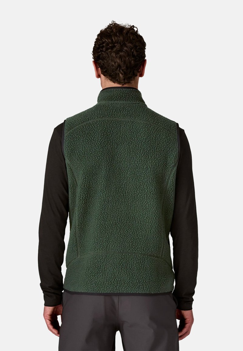 Gilet in pile verde scuro con colletto alto, superficie testurizzata e cuciture a contrasto, indossato sopra una maglietta nera a maniche lunghe.