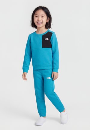 Χαμογελαστό παιδί που φοράει φωτεινό μπλε φούτερ North Face με μαύρο διάκοσμο στο στήθος και αντίστοιχο μπλε παντελόνι φόρμας, περπατώντας με λευκά παπούτσια slip-on.