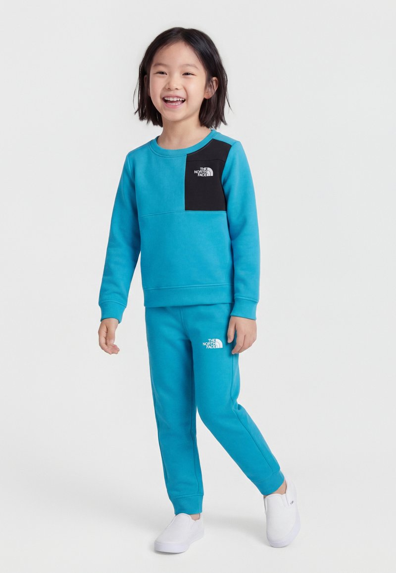 Χαμογελαστό παιδί που φοράει φωτεινό μπλε φούτερ North Face με μαύρο διάκοσμο στο στήθος και αντίστοιχο μπλε παντελόνι φόρμας, περπατώντας με λευκά παπούτσια slip-on.