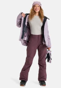Skiovací oblečení s světle fialovou bundou, šedým topem a tmavě fialovými kalhotami. Mezi funkce patří zipy, kapsy a černé rukavice.