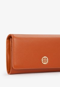 Portafoglio in pelle arancione con una texture liscia, forma rettangolare e un accento circolare dorato. Presenta un design pieghevole e bordi cuciti.