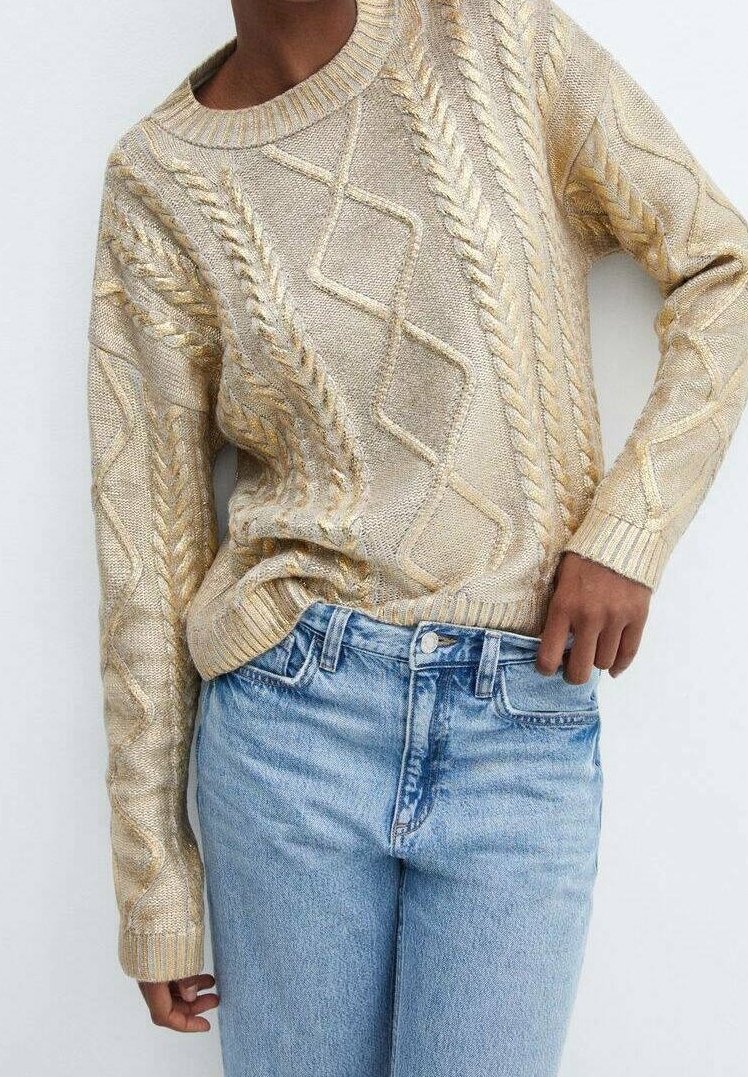 Personne portant un pull beige en maille torsadée rentré dans un jean bleu clair taille haute, debout devant un fond blanc uni.
