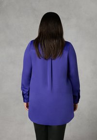 Långärmad skjorta i livfullt lila, med en jämn textur, knappmanschetter och rundad fåll, sedd bakifrån.