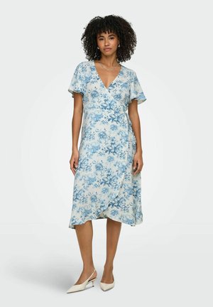 Donna che indossa un vestito avvolgente a fiori azzurro chiaro con maniche corte e tacchi slingback bianchi con punta appuntita, in piedi su uno sfondo semplice.