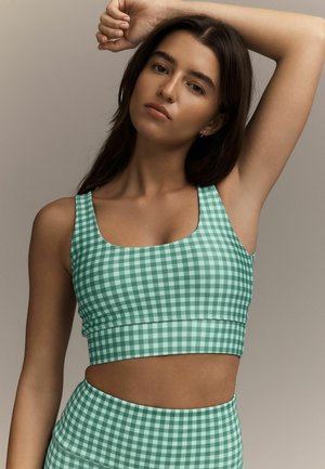 Woman with long dark hair wearing green-and-white checkered crop top and matching high-waisted bottoms, raising one arm above her head.