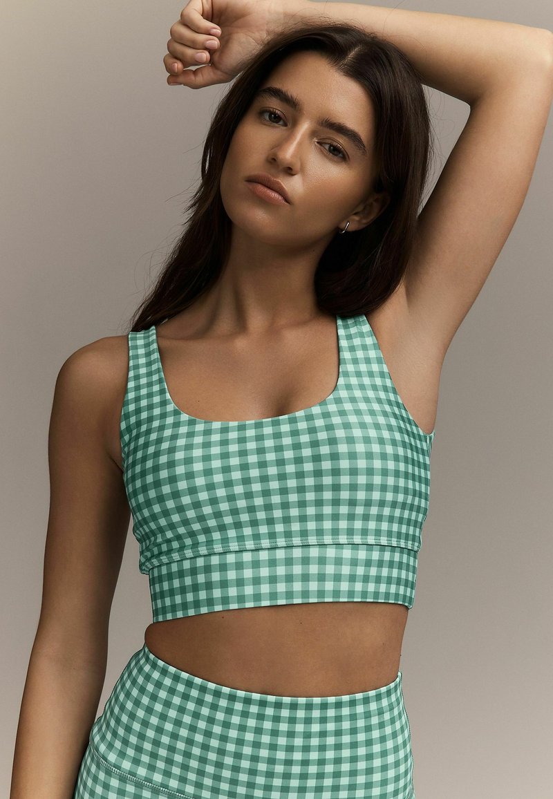 Femme aux longs cheveux foncés portant un crop top à carreaux verts et blancs et un bas taille haute assorti, levant un bras au-dessus de la tête.