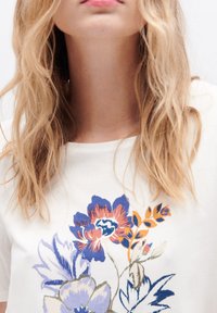 Weißes Baumwoll-T-Shirt mit bunten Blumenstickereien in Blau-, Rot-, Lila- und Orangetönen. Kurze Ärmel, Rundhalsausschnitt.