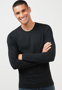 Sort langærmet t-shirt lavet af blødt stof, med rund halsudskæring og en tætsiddende pasform, båret af en smilende person.