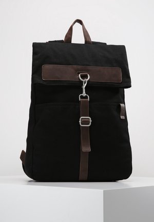 Sac à dos en toile noire avec un rabat en cuir marron. Il comporte des accessoires en métal, une poche avant et des bretelles réglables pour un transport confortable.