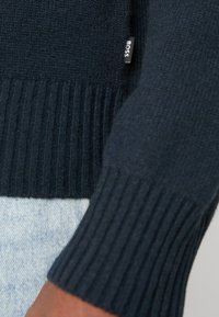 Maglione in maglia navy con polsini a coste. Presenta una texture sottile e una piccola etichetta con il marchio cucita di lato. Primo piano del dettaglio della manica.