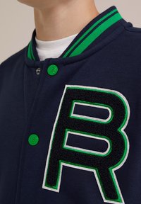 Navyblå varsityjacka med krage i gröna och vita ränder, gröna knappar och ett svart texturerat bokstav "B" accentuerad i grönt och vitt.