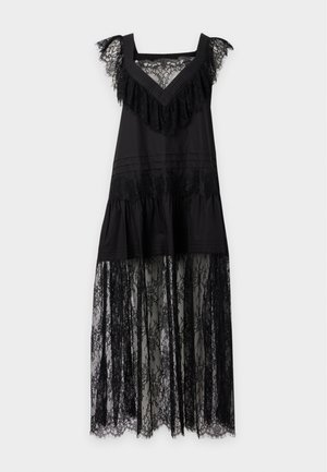 Femme portant une robe noire sans manches avec des détails en dentelle, une jupe inférieure transparente et des ballerines noires, debout devant un fond uni clair.
