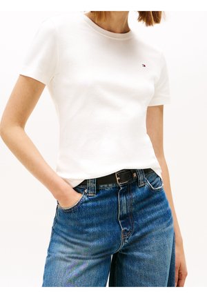 Vrouw draagt een wit T-shirt met korte mouwen en een klein logo op de borst, ingestopt in een high-waist blauwe spijkerbroek met zwarte riem, hand in de zak.