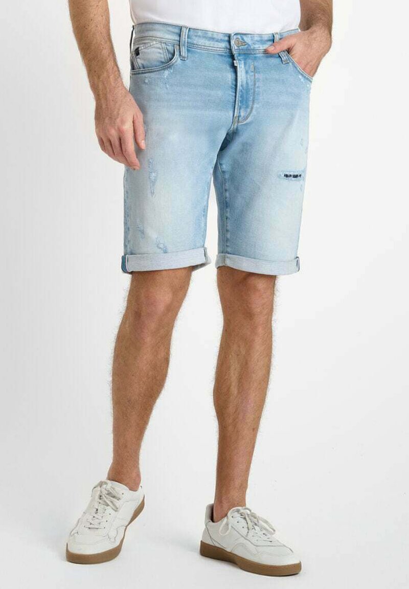 Pantalones cortos de mezclilla azul claro con puños doblados, que presentan un diseño desgastado y detalles desgastados. Combinados con zapatillas blancas.