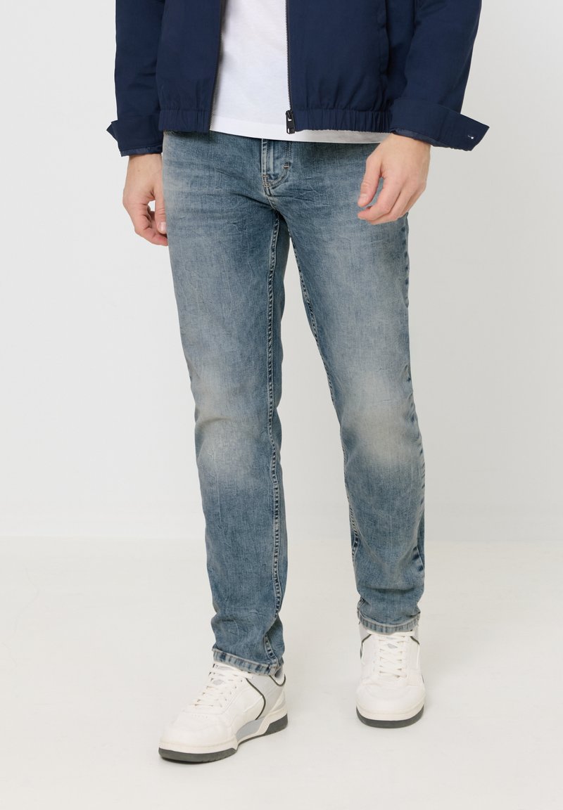Homme portant un jean bleu délavé, des baskets blanches, une chemise blanche et une veste zippée bleu marine, debout devant un fond clair uni.