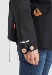 Veste noire avec des accents en cuir beige, des fermetures à boutons et le mot "FORAFEW" brodé. Le tissu semble lisse avec un design structuré.