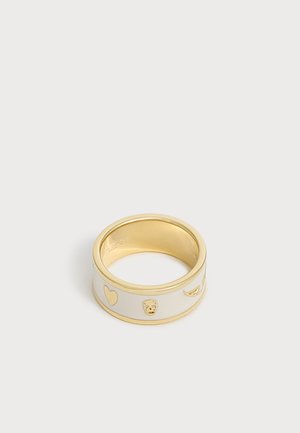 Bague large en or et blanc ornée de symboles en or en forme de cœur, de crâne et d'aile sur un fond clair et uni.