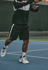 Idrottare i en svart och vit träningsoverall, svarta shorts och vita tennisskor med gula detaljer, hållande ett tennisslag på en tennisbana.