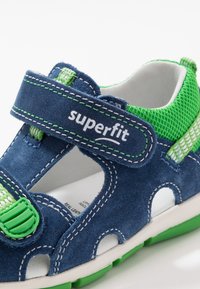 Sininen mokkatossu, jossa on vihreät verkko-yksityiskohdat, tarrasuljenta ja avoimet leikkaukset tuuletusta varten. Varustettu valkoisella tikkaamalla ja kumipohjalla.