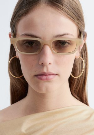 Jeune femme aux cheveux bruns et lisses portant des lunettes de soleil rectangulaires beige, de grandes boucles d’oreilles dorées en forme d’anneaux et un top beige à épaules dénudées.