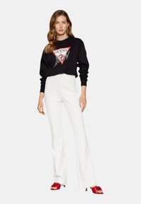 Sudadera negra con logo y gráfico, combinada con pantalones acampanados blancos y sandalias de tacón rojas, ofreciendo un contraste en colores y estilos.