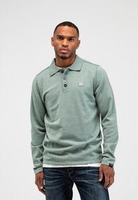 Langarm-Poloshirt aus hellgrünem Stoff, mit Kragen, einer Drei-Knopf-Leiste und einem quadratischen Logo-Patch auf der Brust.