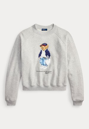 Felpa grigio chiaro con grafica di un orso che indossa una giacca blu navy, maglione bianco, jeans, sneakers, cappellino e porta una borsa bianca.