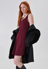 Bordeaux mouwloze jurk met een aansluitend ontwerp. Zwarte jas met nepbontaccenten over de schouders gedrapeerd. Gladde stoftextuur en een knielange snit.