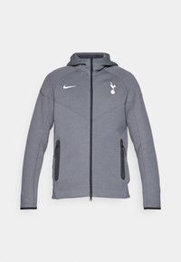 Grå Nike hoodie med fuld frontlynlås, to sidelommer og hætte. Har Tottenham Hotspur-logoet og slanke, moderne sømme.