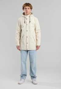 Chaqueta impermeable de color crema con capucha, cordones ajustables, botones de presión y bolsillos laterales, llevada sobre jeans de un azul claro.