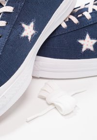 Modré plátěné tenisky s bílou gumovou podrážkou, zdobené růžovými puntíkovanými hvězdičkami a bílými tkaničkami. Tkaničky jsou zobrazeny stočené vedle bot.