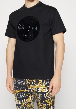 Mężczyzna ubrany w czarną krótką koszulkę z podniesionym czarnym okrągłym logo oraz czarne spodnie z ozdobnym, złoto-białym, barokowym wzorem.