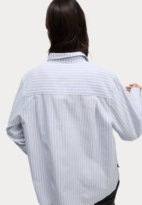 Camisa de rayas en colores azul claro y blanco, con cuello y un corte holgado con hombros caídos. Textura de tela suave.