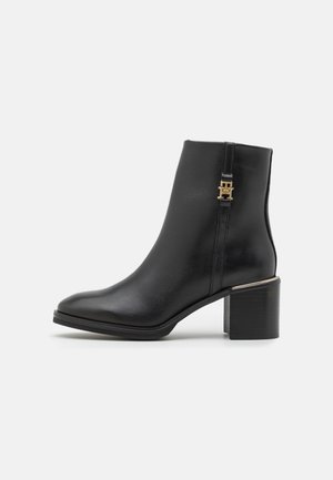 Bottine en cuir noir avec un bout légèrement pointu, un accent en métal doré près de la fermeture éclair et un large talon noir. Texture lisse sur l'ensemble.