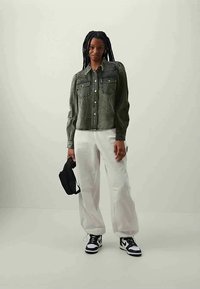 Green denim jacket with two chest pockets, button closure, and long sleeves; paired with loose white pants and black sneakers.