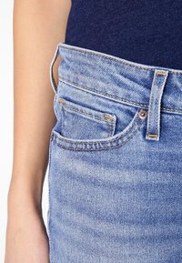 Jeans de mezclilla azul claro con un diseño ajustado, que presentan un pequeño bolsillo frontal, costuras contrastantes y un detalle de botón de metal en la cintura.
