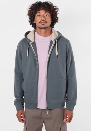 Homme aux cheveux bouclés portant un sweat à capuche gris zippé par-dessus un t-shirt violet clair et un pantalon cargo marron, debout devant un fond clair uni.