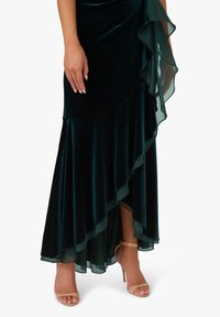 Smaragdově zelené sametové šaty s vrstveným, volánkovým lemem. Průsvitné akcenty zvýrazňují strukturu. Průhledné tenké sandály na podpatku doplňují outfit.