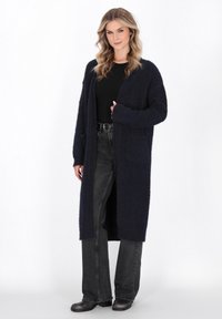 Marinefarbener langer Cardigan mit weicher, flauschiger Textur. Offene Front, lässige Passform, lange Ärmel und frontale Taschen, kombiniert mit dunkelblauen Jeans und Stiefeln.