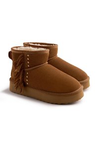 Botas de tobillo de ante marrón con suave forro de piel blanca, flecos decorativos en los lados y suelas de goma texturizadas, mostradas en vista lateral en ángulo.