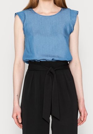 Kobieta w bezrękawniku z jasnoniebieskiego denimu, wsadzonym w spodnie z wysokim stanem w kolorze czarnym, z przewiązanym tkaninowym paskiem w talii.