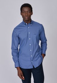 Chemise bleu à boutons, avec un micro-motif, manches longues et col classique. Présente des boutons blancs et un petit logo sur la poitrine.