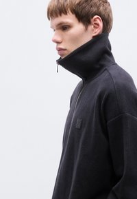 Sweat à capuche noir zippé avec col montant, texture douce, présentant un patch logo subtil sur la poitrine et un design épuré et minimaliste.